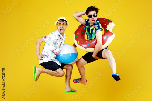 Fototapeta Naklejka Na Ścianę i Meble -  Two young Asian guys in summer casual with hat and sunglasses jumping up high with funny gesture with beach ball and ring float