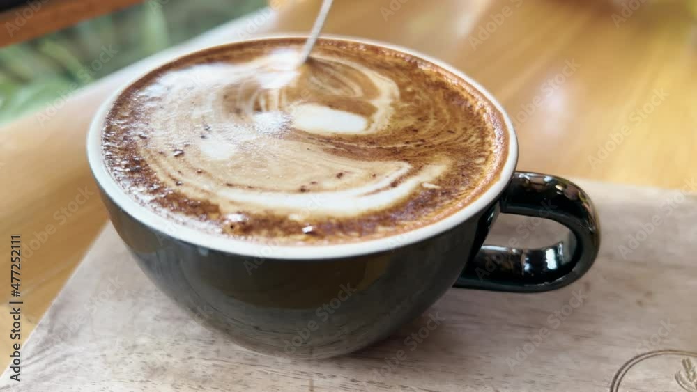 Close-up of fragrant cappuccino coffee with a beautiful latte art pattern in form of a leaf. Person is stirring with an iron spoon a delicious coffee drink standing on a wooden table in a restaurant. Close-up of fragrant cappuccino coffee with a beautiful latte art pattern in form of a leaf. Person is stirring with an iron spoon a delicious coffee drink standing on a wooden table in a restaurant.