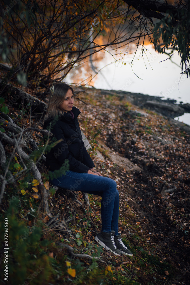 Fototapeta premium Shining young woman sitting on tree roots in forest, smiling, looking away and posing near river with tree in background, wearing blue jeans, black jacket and grey sneakers. Autumn walk in forest.