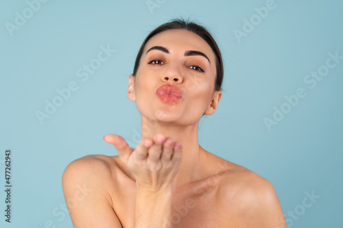 Close beauty portrait of topless woman with perfect skin and natural make-up, plump nude lips, on a blue background, sends a kiss