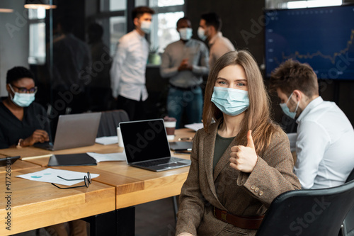 Businesswoman in mask sitting at desk showing thumbs up gesture
