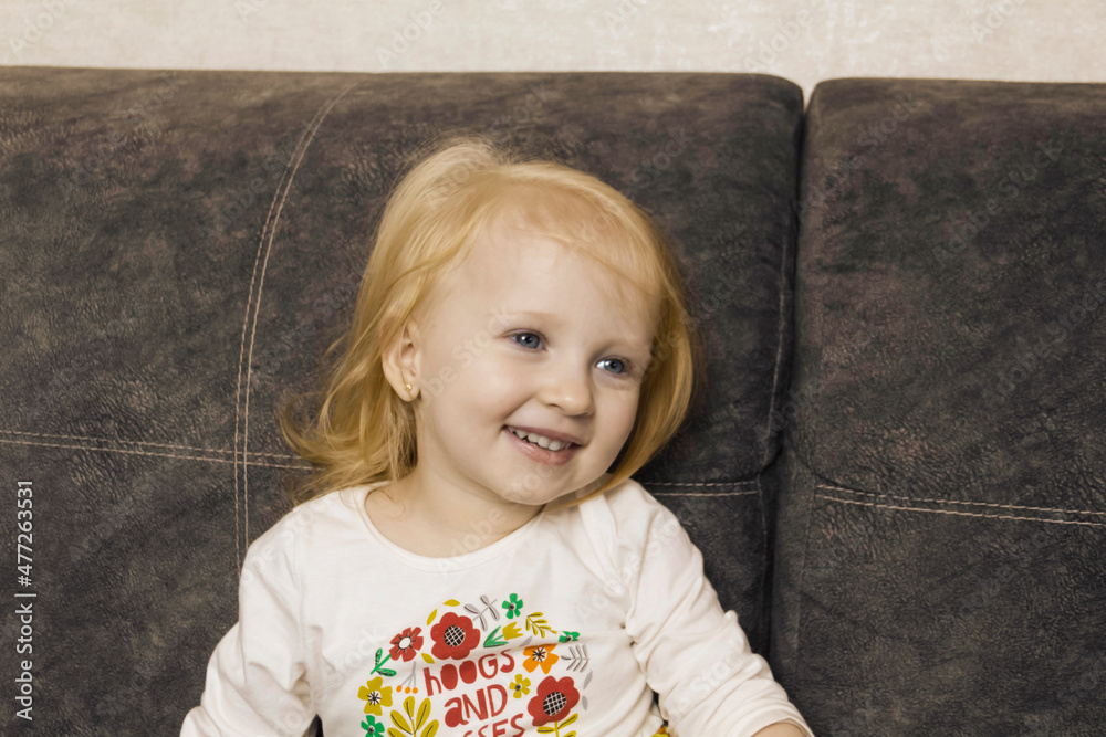Naklejka premium A small, red-haired girl is laughing at home on a gray sofa with purple pillows and a yellow background.
