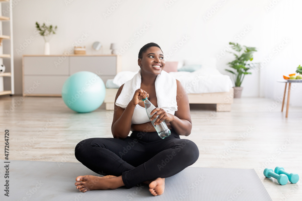 Plump black lady in sportswear sitting on sports mat with bottle of clear water at home, full length Plump black lady in sportswear sitting on sports mat with bottle of clear water at home, full length