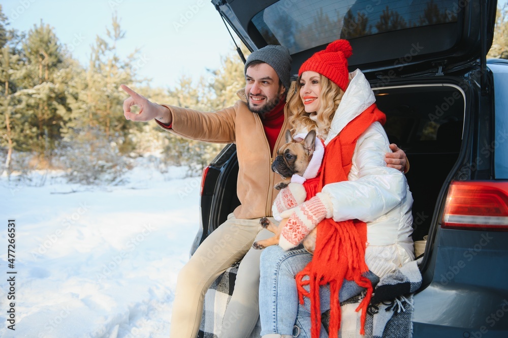 Smiling couple with dog sitting in open SUV car trunk in snowy forest. Enjoying each other in active winter holidays.