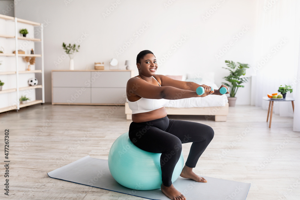 Plus size black woman working out with dumbbells on fitness ball at home, empty space Plus size black woman working out with dumbbells on fitness ball at home, empty space