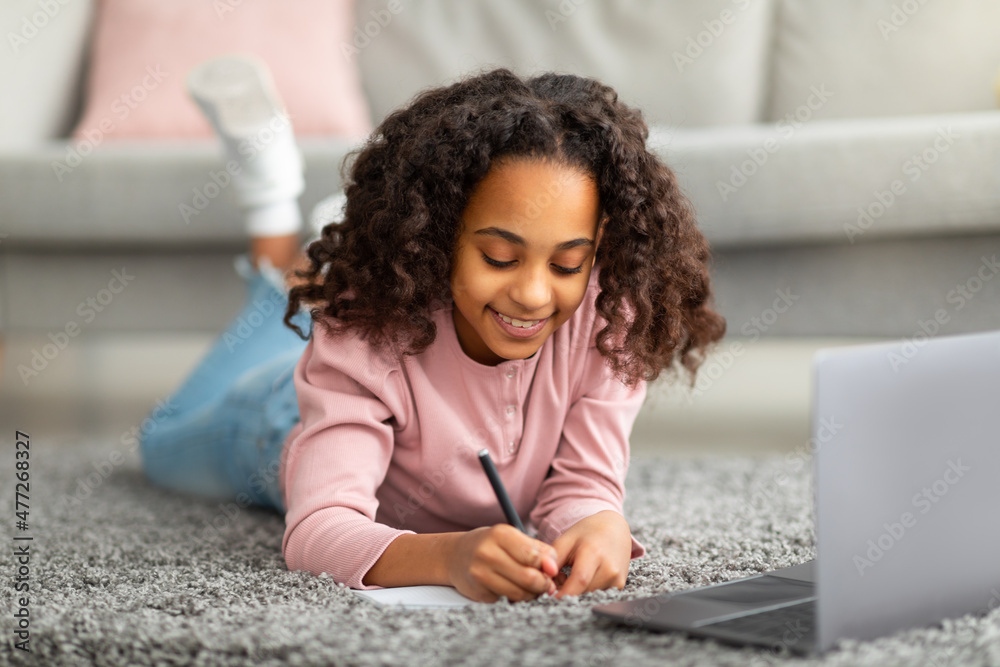 Doing homework. Happy african american girl studying online from home taking notes in front of laptop, lying on carpet Doing homework. Happy african american girl studying online from home taking notes in front of laptop, lying on carpet