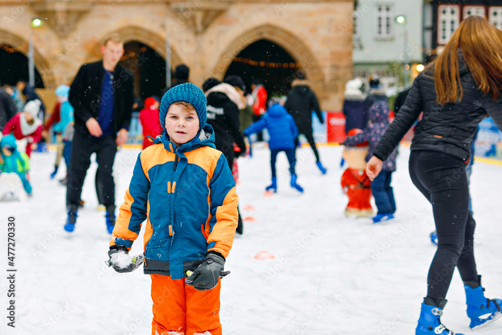 Happy little kid boy in colorful warm clothes skating on a rink of Christmas market or fair. Healthy child having fun on ice skate. Lot of people celebrating holiday and having active winter leisure Happy little kid boy in colorful warm clothes skating on a rink of Christmas market or fair. Healthy child having fun on ice skate. Lot of people celebrating holiday and having active winter leisure