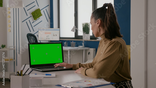 Woman sitting at desk with green screen on laptop, working with chroma key template and isolated mock up background. Person using computer with chroma-key mockup copy space on display.