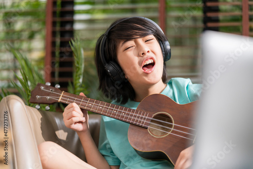 asian child boy sing lound while learn online ukulele study course with fun and exiting at home,online study stay home concept asian child playing ukelele and sing in living room at home photo