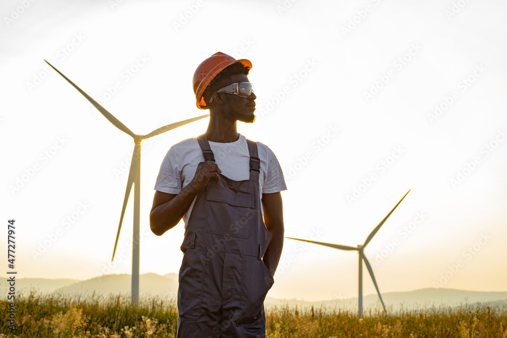 African american man standing on field with huge windmills during summer sunset. Professional technician wearing grey overalls, orange helmet and safety glasses. African american man standing on field with huge windmills during summer sunset. Professional technician wearing grey overalls, orange helmet and safety glasses.