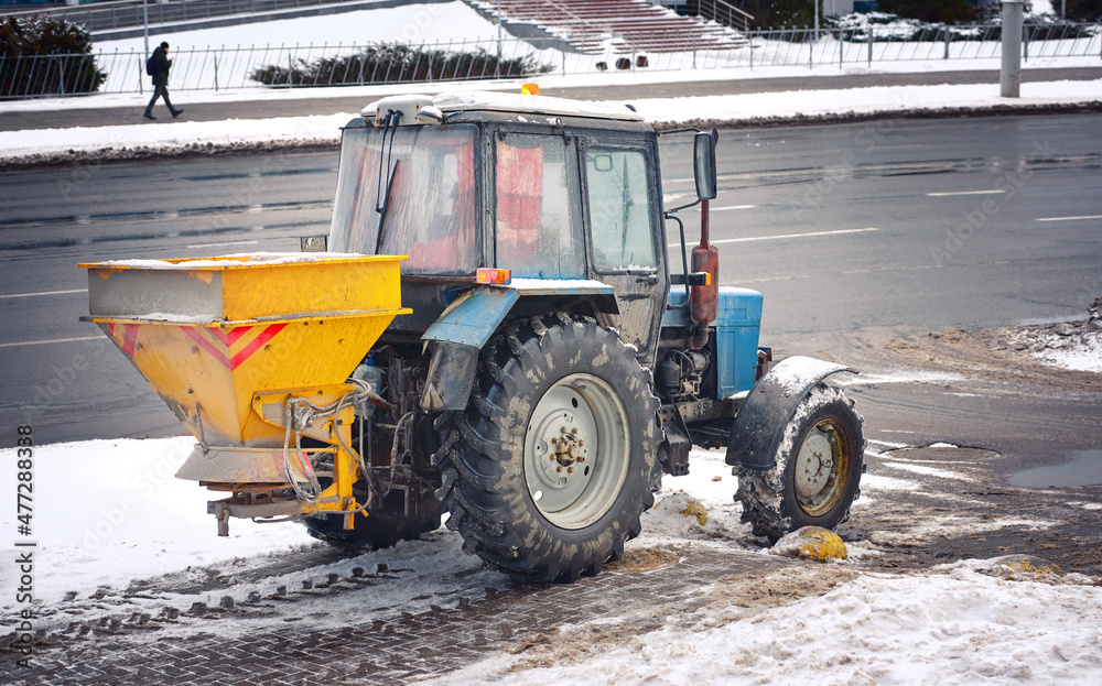 Tractor with mounted salt spreader driving on sidewalk, snow removal service operating in winter. Snow and ice removal service spread rock salt on paving slabs. Municipal workers clearing winter road Tractor with mounted salt spreader driving on sidewalk, snow removal service operating in winter. Snow and ice removal service spread rock salt on paving slabs. Municipal workers clearing winter road