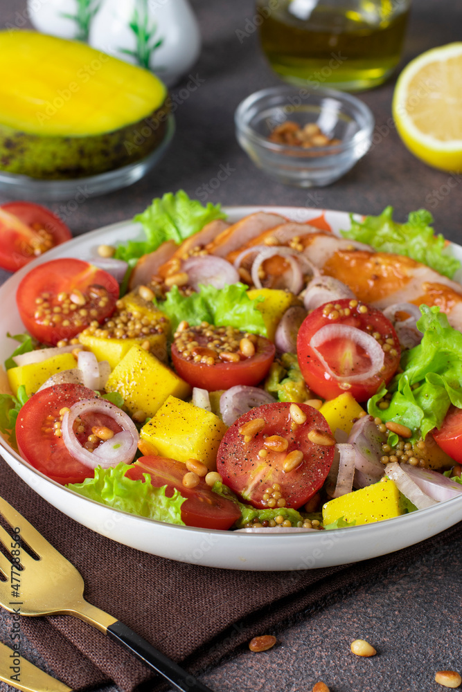 Salad with mango, smoked chicken and cherry tomatoes served with green lettuce leaves in a plate on a brown background