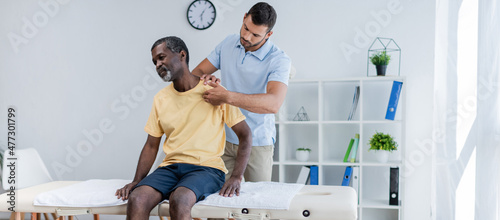 middle aged african american man sitting on massage table while rehabilitologist examining his shoulder, banner photo