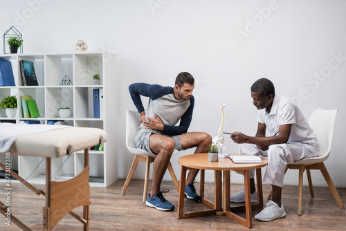 mature african american doctor pointing at spine model near patient suffering from back pain