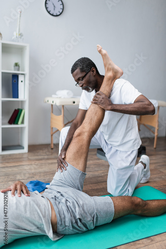 african african rehabilitologist stretching leg of man lying on fitness mat in rehabilitation center