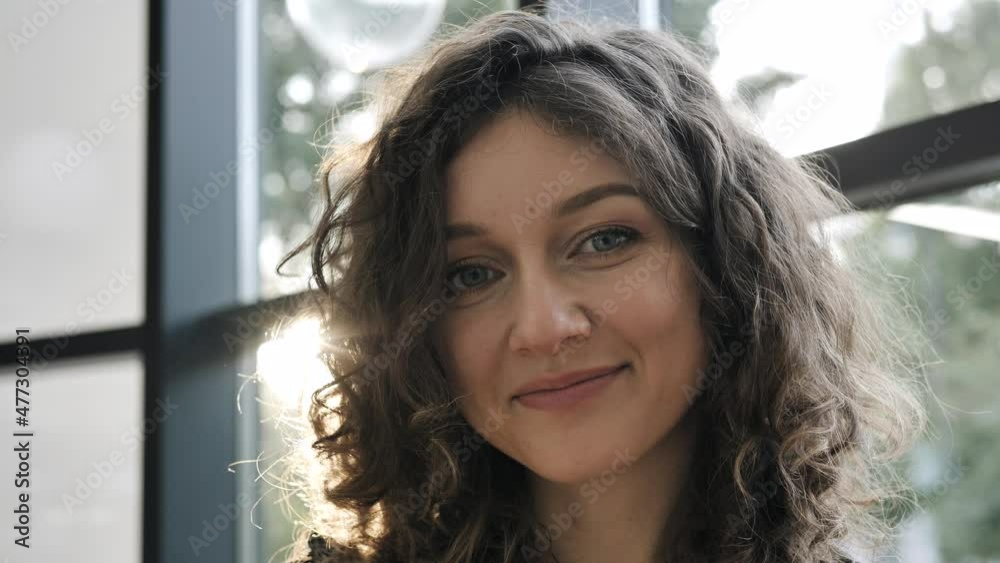 Curly haired young woman with light makeup smiles and poses looking into camera bending head against window at back sunlight Curly haired young woman with light makeup smiles and poses looking into camera bending head against window at back sunlight