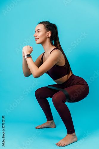 beautiful sportive brunette woman squats with elastic band on blue background. sports exercises. isolated.
