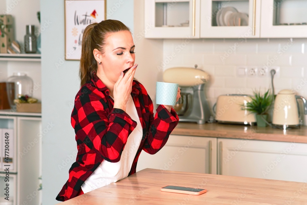 Tired, sweet woman holding a mug of coffee in the kitchen at home, yawns. Girl, the female did not get enough sleep Tired, sweet woman holding a mug of coffee in the kitchen at home, yawns. Girl, the female did not get enough sleep