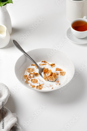 Bowl of wholebread cornflakes with milk on a white table with a cup of tea. Breakfast time. White photography.