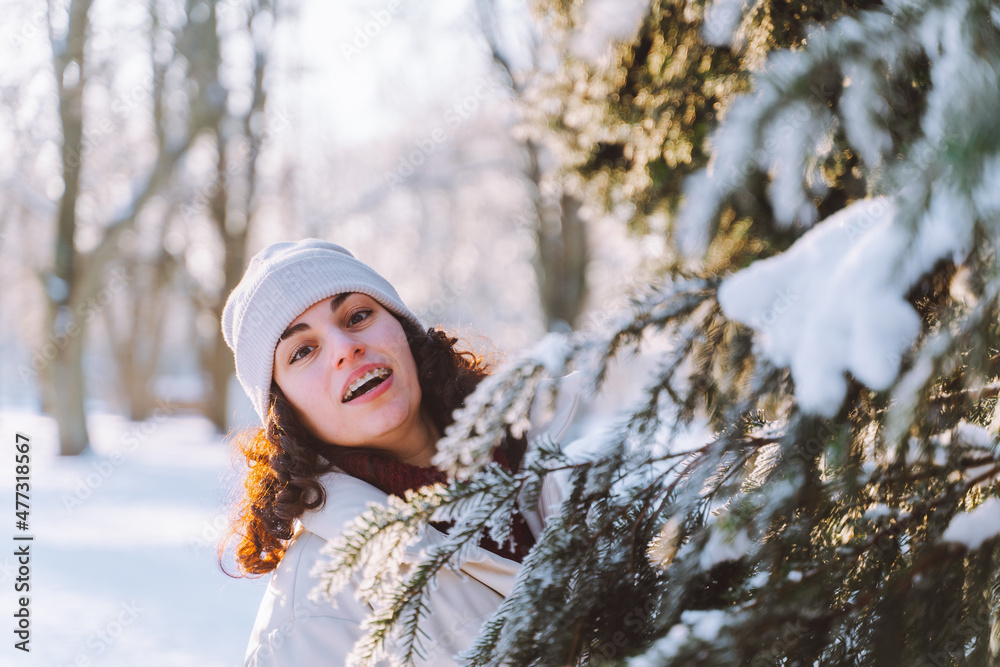 Young curly smiling woman walk in winter park and hide behind snow-covered Christmas tree. Positive emotions, freshness, holidays Young curly smiling woman walk in winter park and hide behind snow-covered Christmas tree. Positive emotions, freshness, holidays