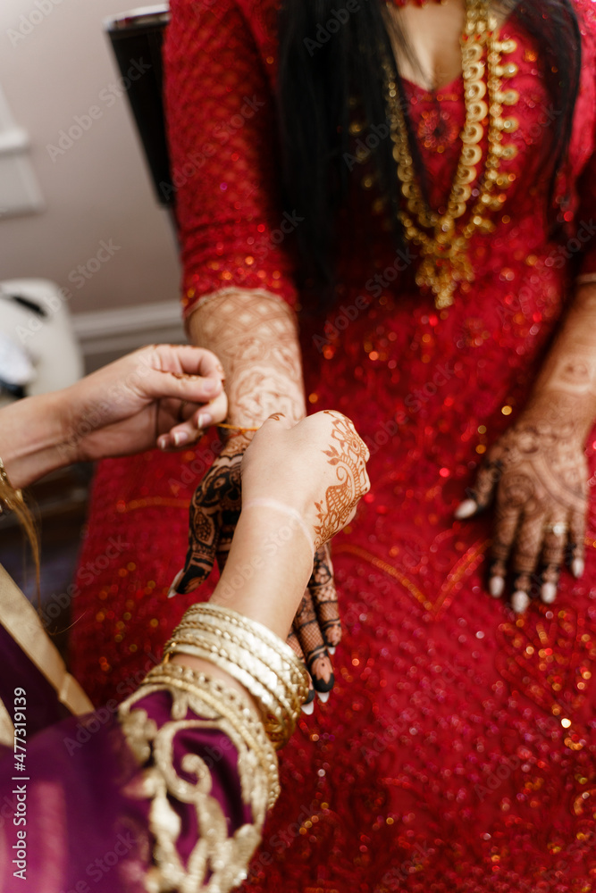 Bridesmaid helps Hindu bride to put bangles and bracelet Jewelry Woman in pink sari puts golden bracelet with bells on bride's hand painted with mehndi Wedding preparations Hindu Bride in Red dress