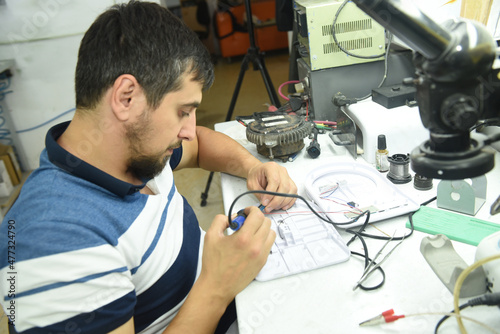 A young man at a desk in a workshop laboratory repairing a chip with an electric soldering iron