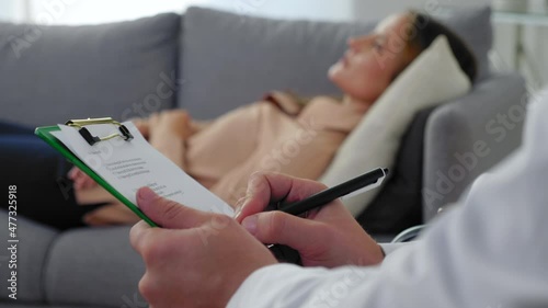 Close up man psychotherapist taking notes on clipboard and listen patient at psychotherapy session. Unfocused adult woman with mental health problem after covid-19 lying on couch talking to therapist
