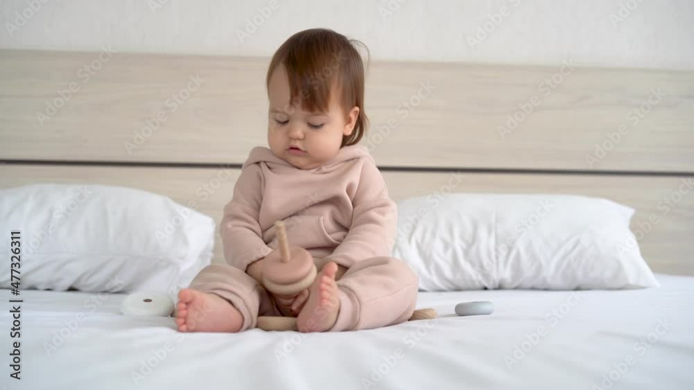 toddler girl sitting on the bed playing with a wooden toy. Children's hands holding wooden blocks of a pyramid. toys teaching children 1 year old. Eco friendly toddler girl sitting on the bed playing with a wooden toy. Children's hands holding wooden blocks of a pyramid. toys teaching children 1 year old. Eco friendly