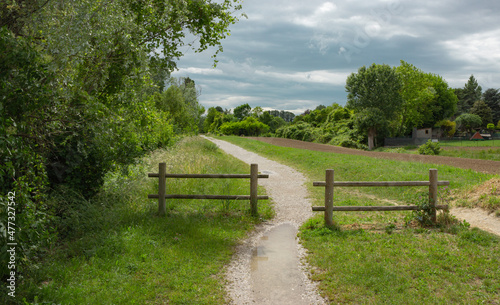 Countryside landscape. White gravel path along the fields with two wooden fences. Puddles on the trail. Cloudy dark sky.