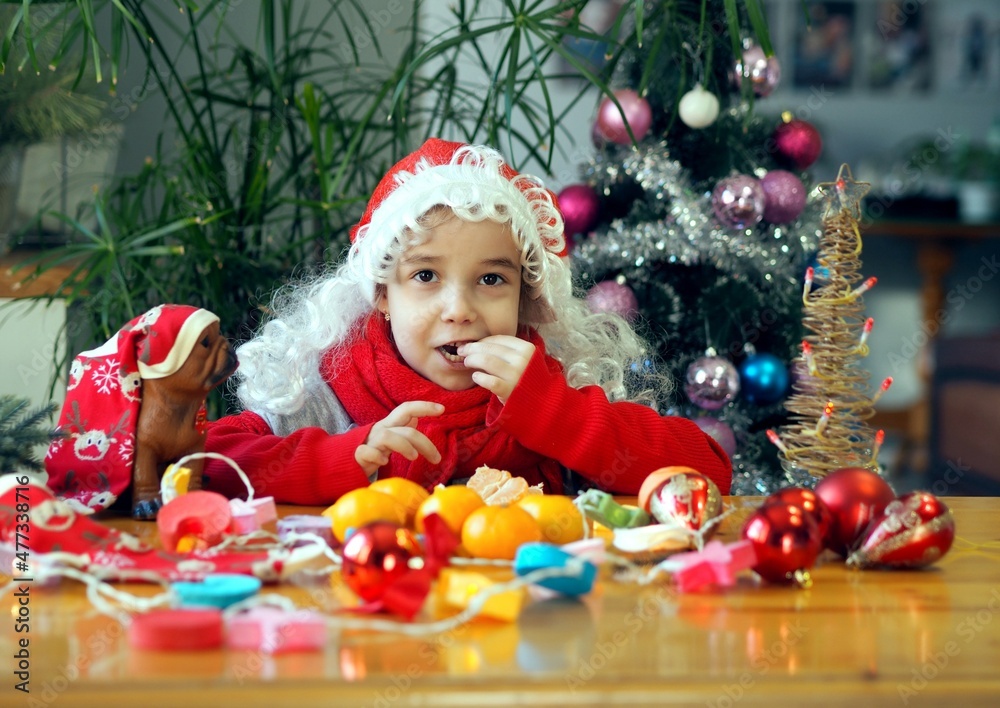 Little girl in Santa Claus hat rejoices and eats tangerines. On the eve of the meeting of Christmas or New Year.
