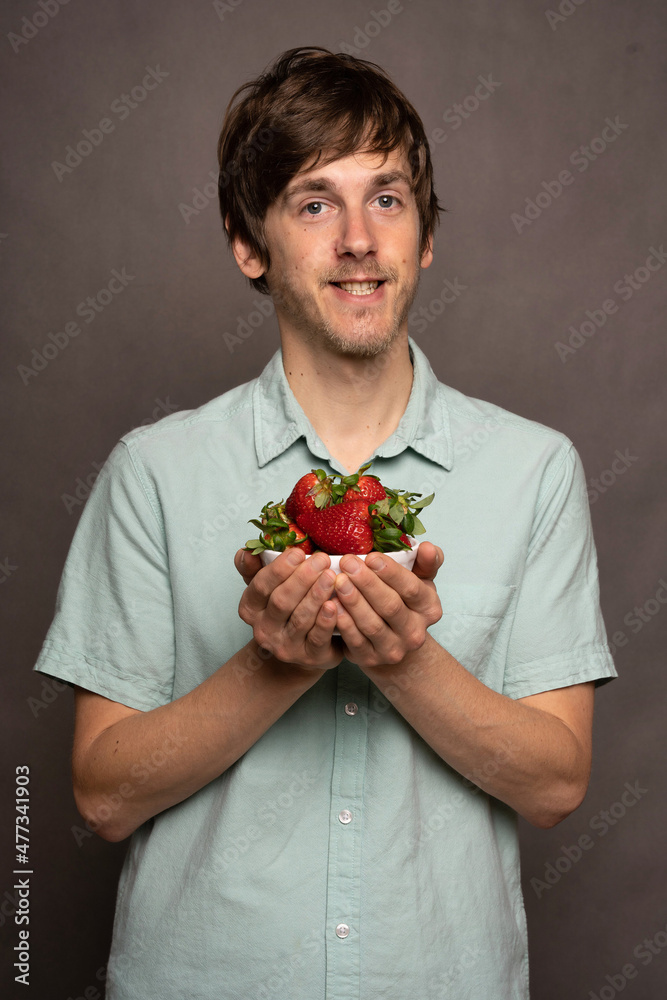Young handsome tall slim white man with brown hair holding strawberries smiling in light blue shirt on grey background