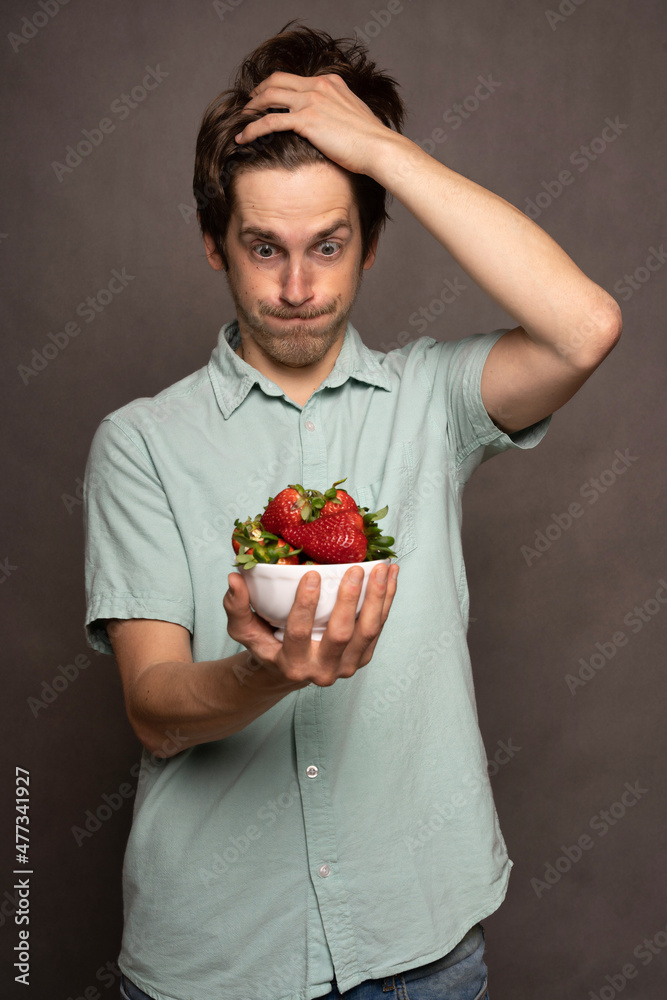 Young handsome tall slim white man with brown hair holding strawberries speechless in light blue shirt on grey background Young handsome tall slim white man with brown hair holding strawberries speechless in light blue shirt on grey background