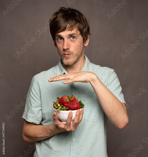 Young handsome tall slim white man with brown hair hovering his hand over strawberries in light blue shirt on grey background