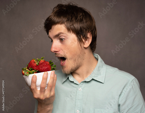 Young handsome tall slim white man with brown hair holding strawberries mouth opened in light blue shirt on grey background