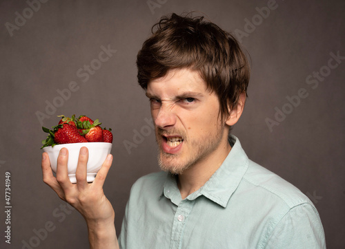 Young handsome tall slim white man with brown hair holding strawberries angrily in light blue shirt on grey background