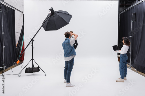 art director holding laptop with blank screen near photographer and model working in photo studio.