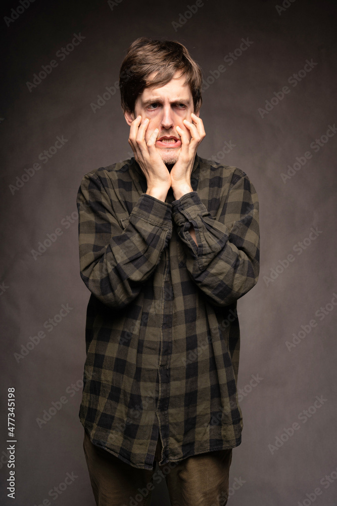 Young handsome tall slim white man with brown hair holding his face in horror with flannel shirt on grey background Young handsome tall slim white man with brown hair holding his face in horror with flannel shirt on grey background