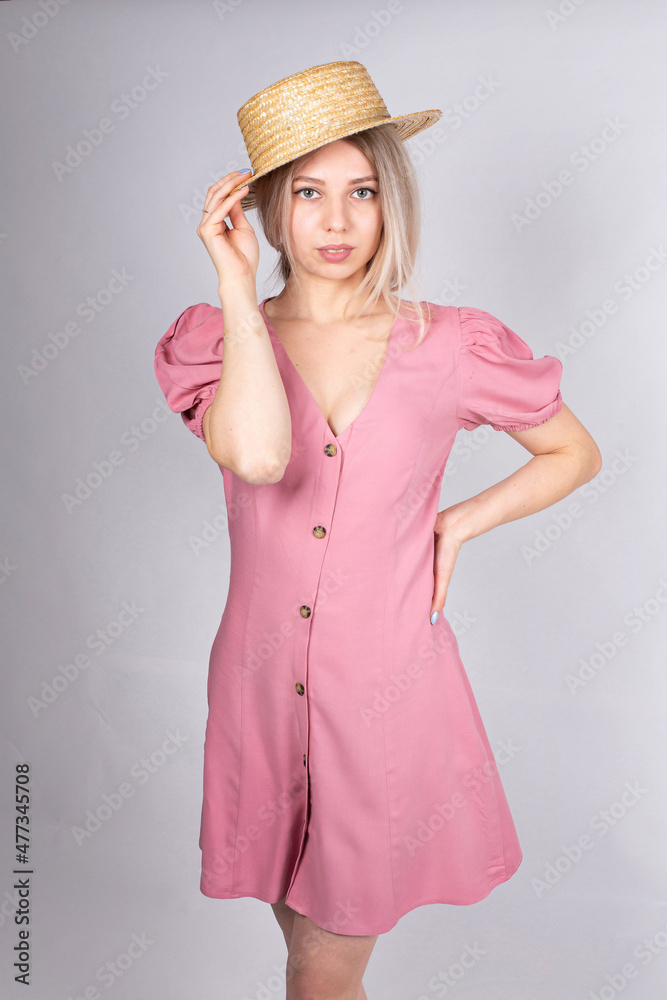 Woman with blonde hair and white skin isolated on grey in pink dress touching her hat by hand Woman with blonde hair and white skin isolated on grey in pink dress touching her hat by hand