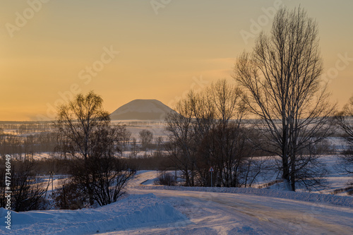 Snow-covered road against the backdrop of a secluded mountain. Everything around is lit by the evening sun.