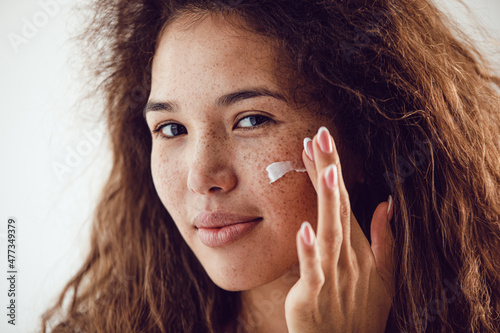 Portrait of woman with curly hair and freckles applying moisturizer to her face.