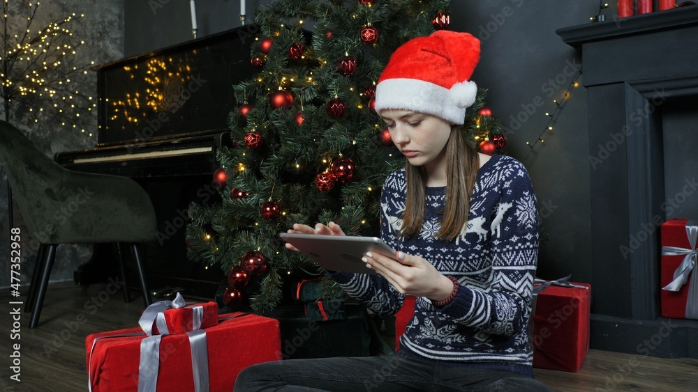 Cute teen girl using tablet near christmas tree, christmas eve, online shopping and entertainment Cute teen girl using tablet near christmas tree, christmas eve, online shopping and entertainment