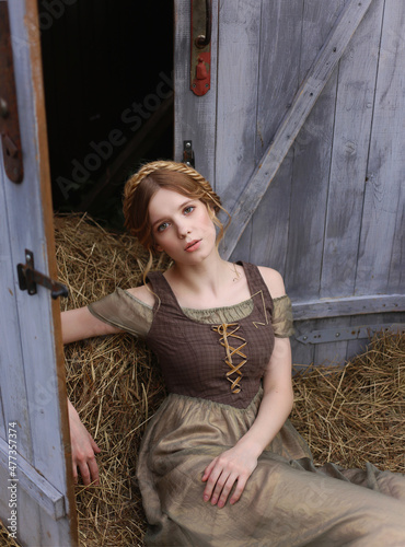 Beautiful young woman with braided hair in simple vintage dress laying on hay near barn doors