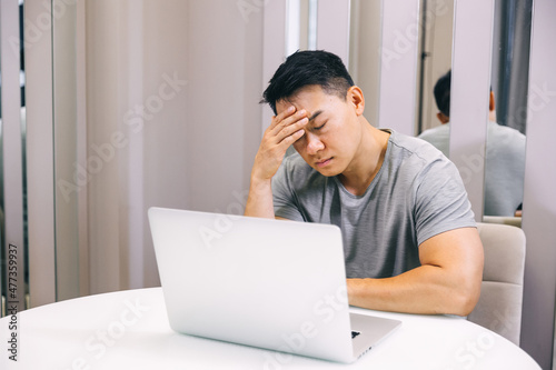 Young tired manager sitting at home with laptop computer having tired look holding his hand on head having pain closing his eyes being sleepy and exhausted. Tiredness