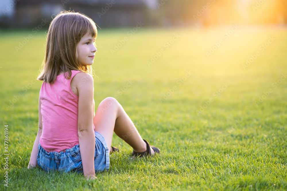 Young happy child girl resting while sitting on green grass lawn on warm summer day Young happy child girl resting while sitting on green grass lawn on warm summer day