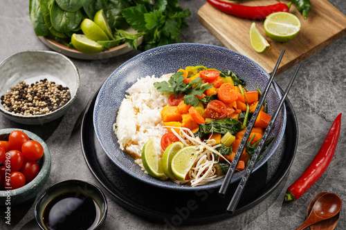 A blue bowl with red thai vegetable curry and basmati rice, fresh lime, cilantro, spinach, chilli peppers and cherry tomatoes
