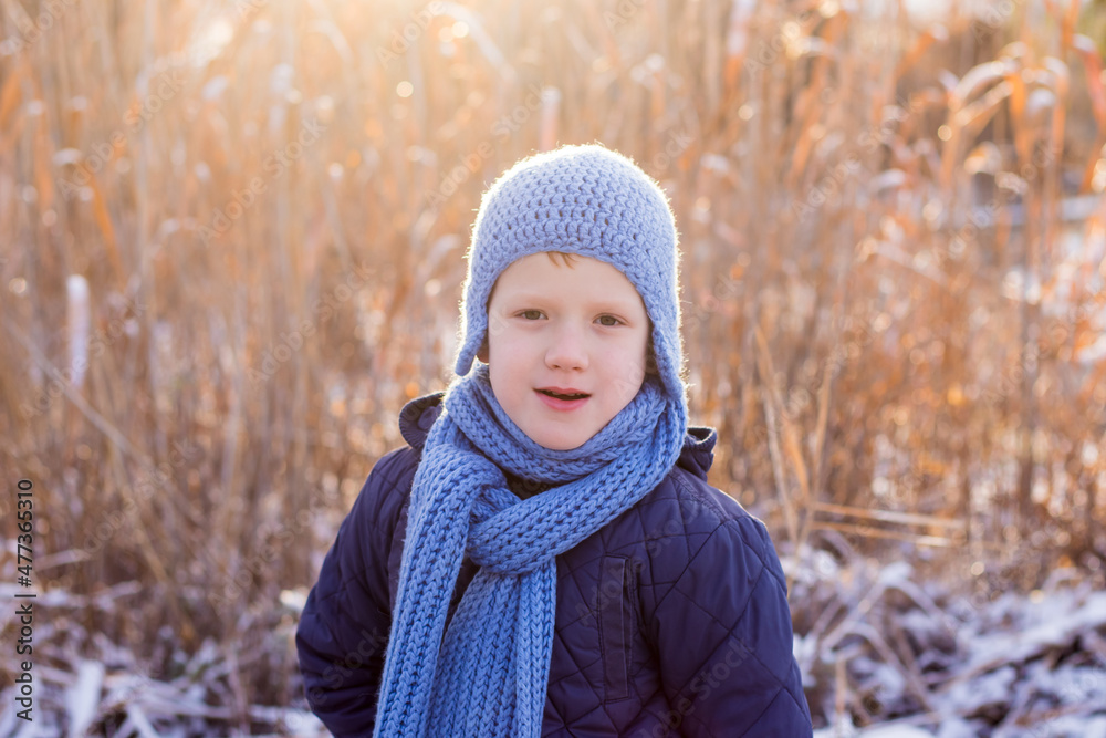 Cute smiling boy wearing bright blue knitted hat, blue jacket, blue scarf outdoors on sunset in a beautiful winter snowy day. Cute smiling boy wearing bright blue knitted hat, blue jacket, blue scarf outdoors on sunset in a beautiful winter snowy day.