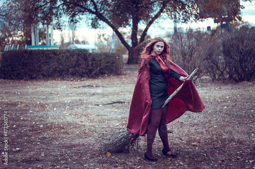 A young beautiful girl in a red cloak with a big broom in her hands, a riddle girl, a witch girl with red hair.