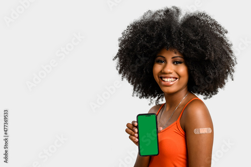 black girl without face shield holds the cell phone close to her arm with the vaccine sticker, fully immunized, chroma key on the cell phone screen, on gray background photo