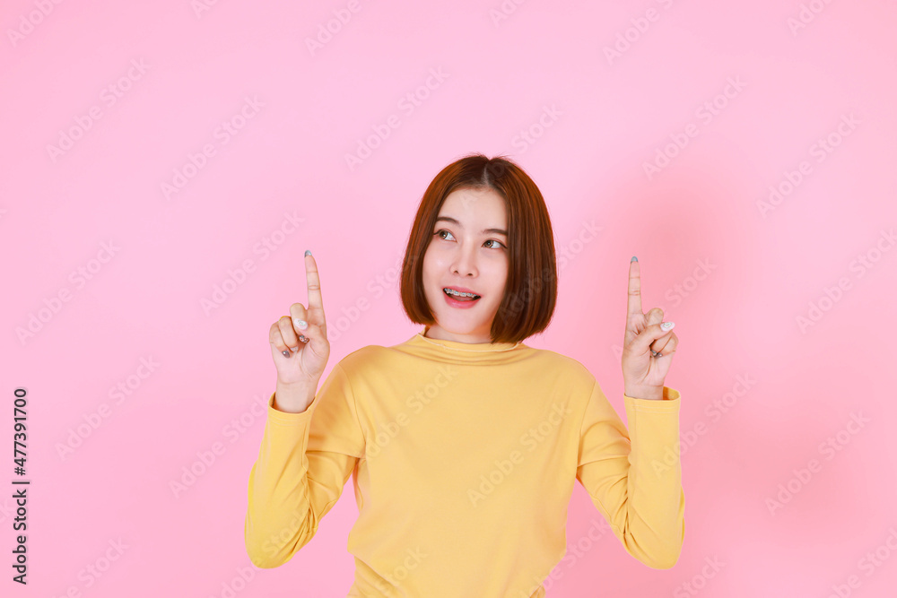 Portrait studio cutout shot of Asian young pretty short hair female model in yellow long sleeve shirt stand smile to look at the camera and pointing finger on pink background. Portrait studio cutout shot of Asian young pretty short hair female model in yellow long sleeve shirt stand smile to look at the camera and pointing finger on pink background.