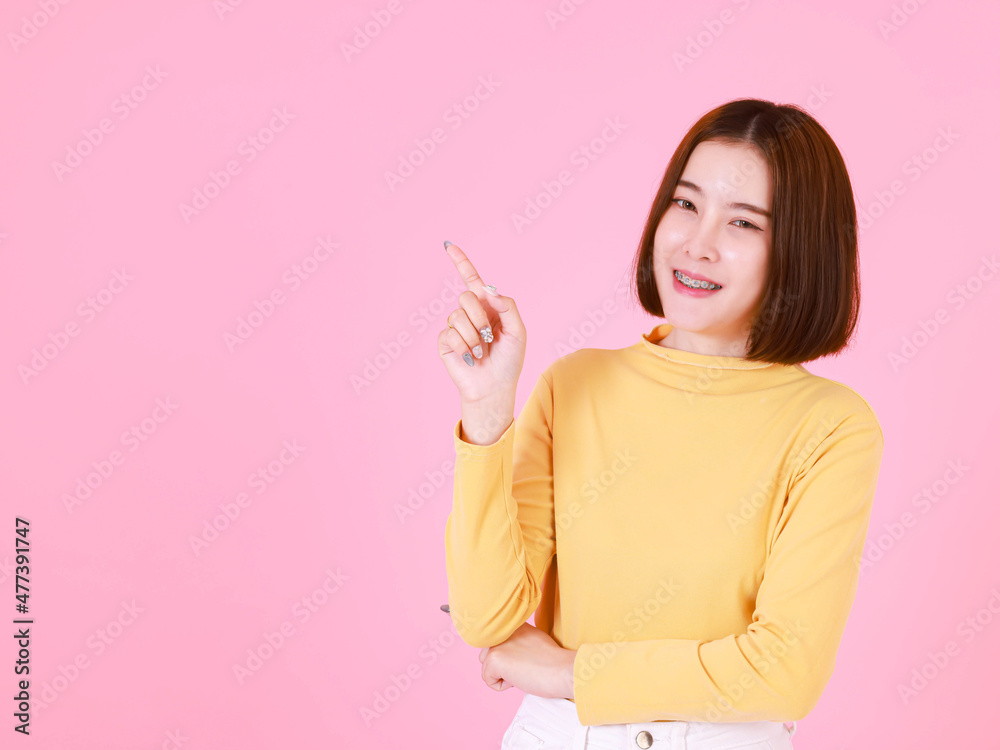 Portrait studio cutout shot of Asian young pretty short hair female model in yellow long sleeve shirt stand smile to look at the camera and pointing finger on pink background. Portrait studio cutout shot of Asian young pretty short hair female model in yellow long sleeve shirt stand smile to look at the camera and pointing finger on pink background.
